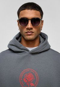 Grijze hoodie met een rode roos graphic, voorzien van een trekkoordkap en een zachte textuur. Ronde, donkere zonnebril maakt de look compleet.