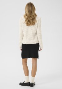 Cremfarbener Strickpullover mit strukturiertem Muster, kombiniert mit einem schwarzen knielangen Rock und schwarzen Slippers, ergänzt durch weiße Socken.