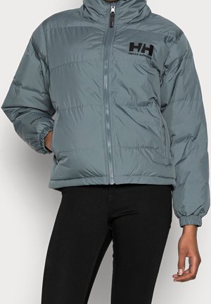Chaqueta acolchada azul grisáceo Helly Hansen con cremallera frontal, puños elásticos y logo en el pecho y la manga, usada sobre pantalones negros.