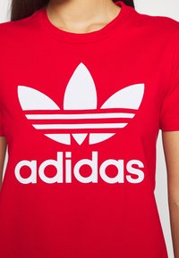 Červené bavlněné tričko s velkým bílým logem Adidas, které má tři paralelní pruhy a listovité designové prvky umístěné na hrudi.