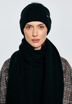 Černá pletená čepice s žebrovanou texturou a malým logem, doplněná tlustou černou šálou z měkké látky; modelka má na sobě kostkovaný kabát.
