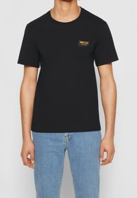 Černé bavlněné tričko s kulatým výstřihem a krátkými rukávy. Obsahuje vyšitý obdélníkový znak s nápisem "MOSCHINO MILANO" ve zlatém písmu.