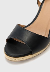 Black leather wedge sandals featuring a closed toe and a wide strap. The sole is textured with an espadrille-style finish.