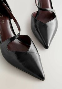 Czarne wysokie obcasy ze skóry z ostrym noskiem, z krzyżującymi się paskami i gładką fakturą, na neutralnym tle.