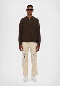 Maglione marrone lavorato a maglia con mezza zip, abbinato a pantaloni beige con intricate decorazioni in argento e sneakers bianche. Occhiali da sole neri.