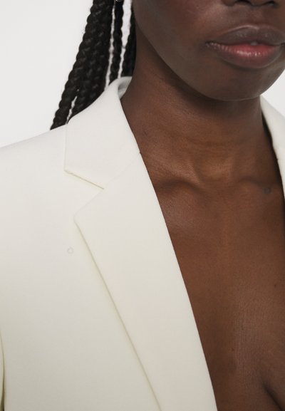 Nærbillede af en person med mørkt fletningehår iført en hvid blazer, der viser deres nakke, læber og en del af brystet.