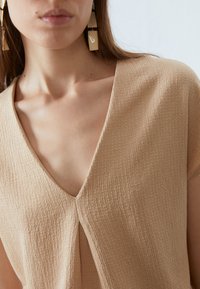 Beige, textuurrijke top met een V-hals en korte mouwen. Opvallende gestikte details in het midden. Vierkante, lichtgekleurde oorbellen met een geometrisch ontwerp.