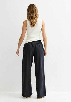 Femme aux longs cheveux ondulés portant un haut blanc sans manches et un pantalon noir ample, debout face à un mur blanc uni.