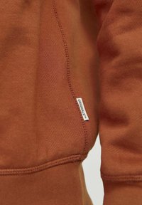 Gros plan sur le tissu d'un sweat-shirt couleur rouille montrant des coutures détaillées et une petite étiquette blanche avec le texte « ORIGINALS™ ».