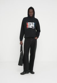 Melnas kapuces džemperis ar grafisku sarkanu, zilu un baltu apdruku, valkāts ar melnām platām biksēm un melnām iešļūcenes, tur tumšu somu.