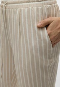 Lettere, stribede beige bukser med en blød tekstur, der har en elastisk talje og sidelommer. De lodrette striber er off-white.