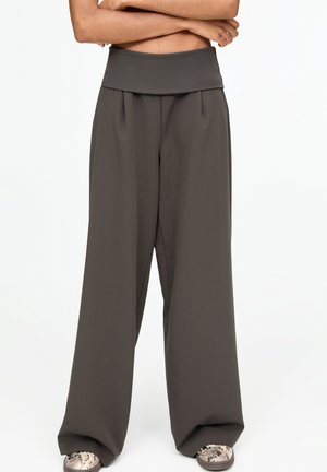 Persona che indossa pantaloni larghi di colore grigio scuro con vita alta, in piedi con le braccia incrociate, abbinati a scarpe con motivo in pelle di serpente.