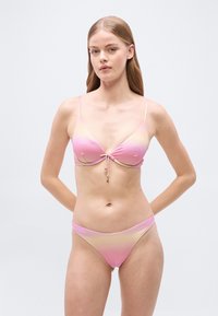 Νεαρή γυναίκα που φοράει ροζ και κίτρινο μπικίνι με διαβάθμιση, στέκεται με τα χέρια πίσω από την πλάτη μπροστά σε ένα απλό φόντο.