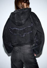 Ženska nosi črno jeans jakno s kapuco in krilato nalepko s srcem na hrbtu, v kombinaciji z usklajenimi visokimi jeans hlačami.