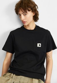 T-shirt noir à manches courtes en coton, doté d'une poche poitrine avec une étiquette carrée. Le design est simple et classique.