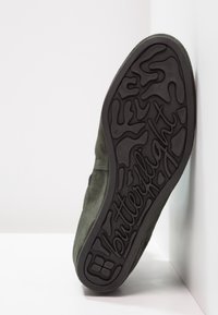 Chaussure en daim vert foncé avec une semelle en caoutchouc noir texturée, présentant un motif distinctif "butterflight" pour une adhérence et une flexibilité.