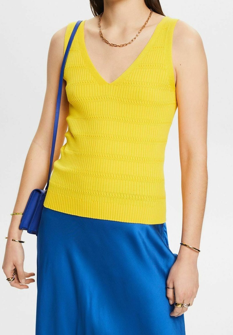 Femme portant un top en maille jaune sans manches avec un col en V, une jupe bleue, des bijoux en or et portant un petit sac à bandoulière bleu.