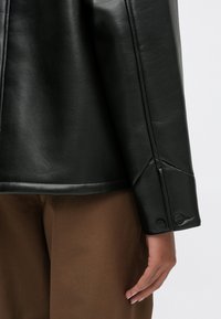 Czarna skórzana kurtka o wyrazistej sylwetce, z zapięciem na guzik przy mankietach i gładką teksturą, w połączeniu z brązowymi materiałowymi spodniami.