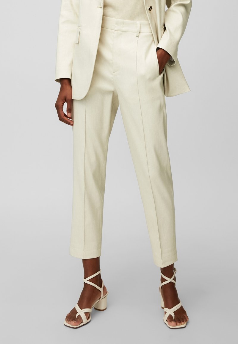 Pantalons beige clair ajustés avec une coupe droite, comprenant des poches latérales et une texture verticale subtile. Associés à des talons beiges à lanières.