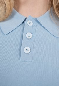 Gros plan sur le col d'un polo en maille bleu clair avec trois boutons ronds, porté par une personne aux cheveux blonds.