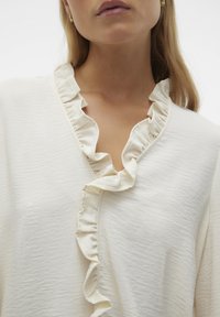 Blouse crème avec un col à volants, tissu doux avec texture, et un design en V. Les volants ajoutent un détail délicat au col.