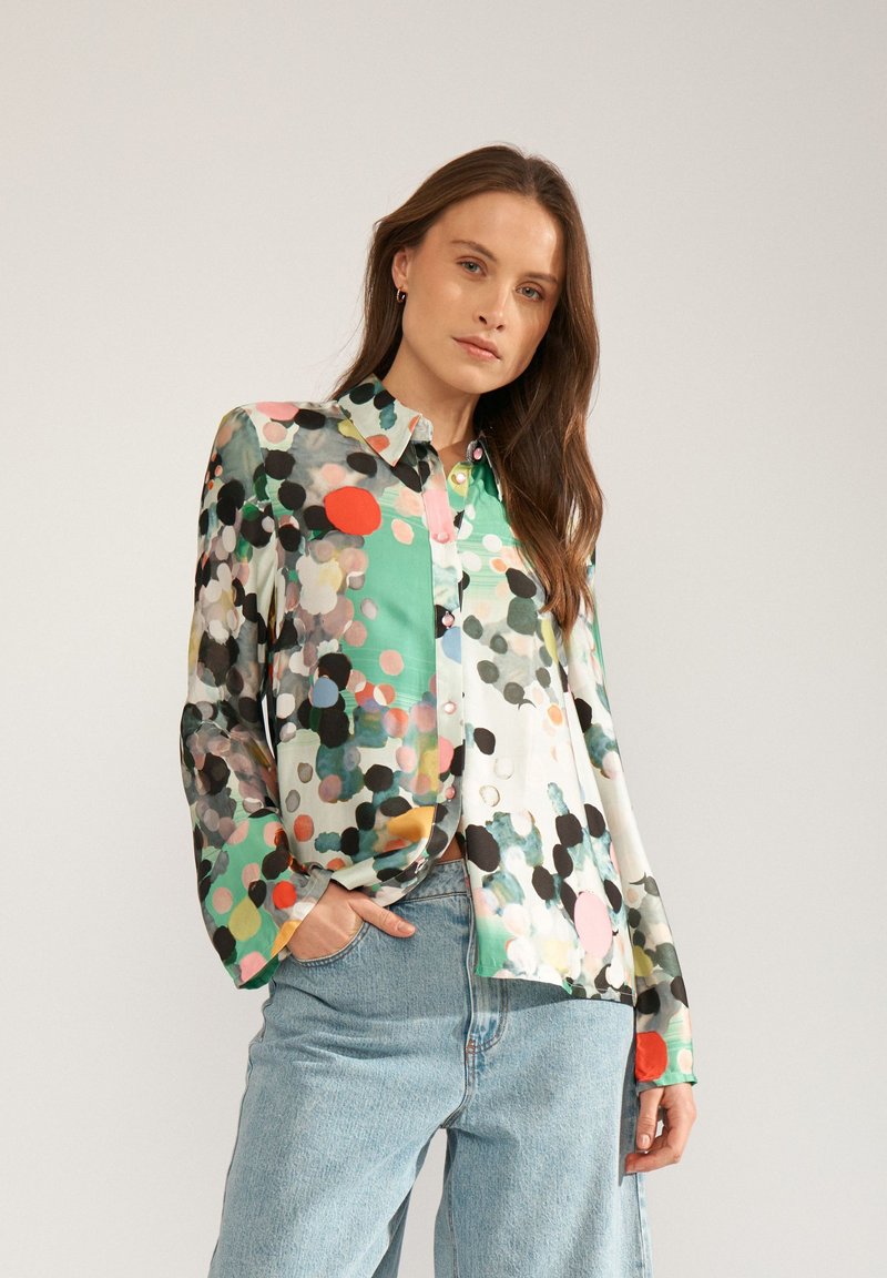 Femme aux longs cheveux bruns portant un chemisier à manches longues aux motifs abstraits et colorés et un jean bleu clair, debout devant un fond gris uni.