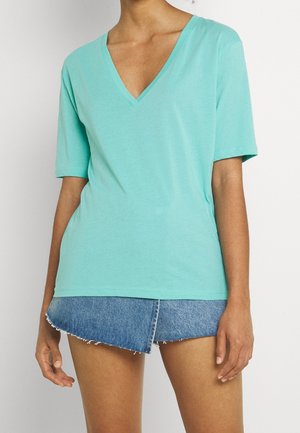 Vrouw draagt een licht turquoise V-hals T-shirt met korte mouwen en een asymmetrische denim minirok met rafelige zomen, gefotografeerd tegen een effen achtergrond.