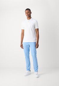 Hvid polo shirt med kortærmer, parret med lyseblå slim-fit bukser. Modellen har hvide sneakers på og står foran en neutral baggrund.
