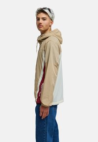 Veste légère à capuche en beige et blanc, avec une bande bordeaux. Confectionnée dans un tissu lisse avec des poignets élastiques et un détail logo sur le devant.