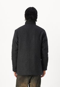 Tmavě šedý vlněný kabát s vysokým límcem, rovný střih a texturovaný povrch. Na švech jsou viditelné stehy, nošený s kalhotami světlé barvy.