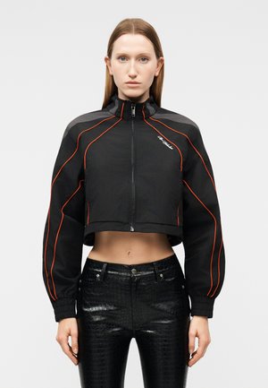 Γυναίκα που φοράει μαύρο κοντό μπουφάν με πορτοκαλί λεπτομέρειες και μαύρο γυαλιστερό παντελόνι ψηλής μέσης με υφή, στέκεται μπροστά σε μία απλή φόντο.