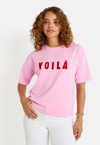 Camiseta rosa de gran tamaño con mangas cortas que presenta la palabra "VOILÀ" en letras rojas y en negrita. Fabricada en tejido suave, con textura lisa.