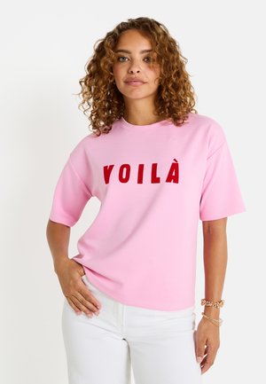 Camiseta rosa de gran tamaño con mangas cortas que presenta la palabra "VOILÀ" en letras rojas y en negrita. Fabricada en tejido suave, con textura lisa.