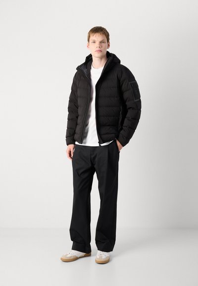 Veste noire matelassée avec capuche, dotée d'une fermeture éclair à l'avant et de poignets élastiques, portée par-dessus une chemise blanche et un pantalon noir ; accompagnée de baskets blanches.