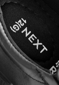 Interior negro de una zapatilla que muestra un forro textil con el texto impreso en blanco "NEXT 12". Material suave y sutiles detalles de costura.