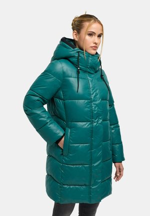 Woman wearing a teal quilted puffer coat with a hood and hands in pockets, facing slightly to the right on a plain background.