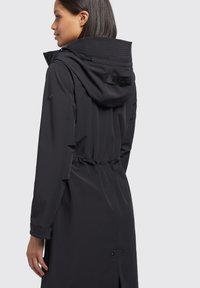 Giacca nera impermeabile con cappuccio regolabile, vita elastica e dettagli con bottoni. Presenta un tessuto texturizzato e linee di design eleganti.