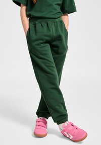 Grüne Sweatpants mit elastischem Bund, tailliertem Beinschnitt und seitlichen Taschen, kombiniert mit rosa Turnschuhen mit Klettverschlüssen und weißen Akzenten.