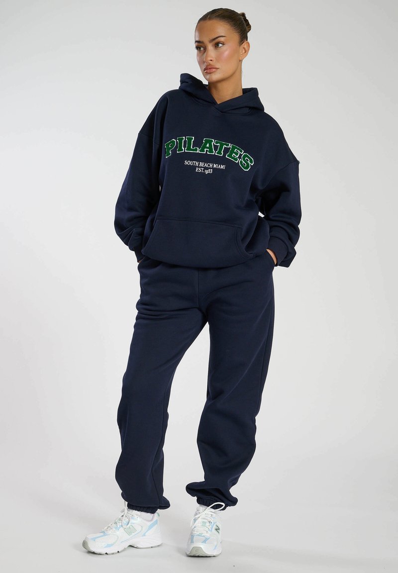 Felpa blu navy con la scritta "PILATES" in verde, abbinata a pantaloni coordinati. Presenta una tasca frontale e polsini a costine. Indossata con sneakers bianche.