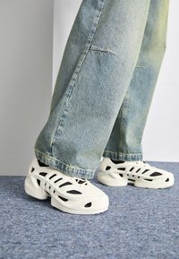 White athletic shoes with cut-out designs and black accents, paired with light blue denim jeans, set against a textured blue carpet.