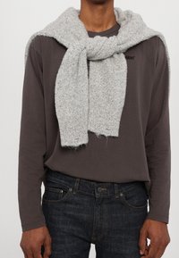 Jasnoszara, dziana szalik ułożony na ciemnej koszuli z długim rękawem, z widoczną teksturą i strzępionymi końcami. Ciemne dżinsy z kieszeniami i szwami.