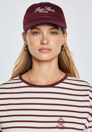 Tamsiai raudonas beisbolo kepuraitė su baltais siūlais išsiuvinėtu užrašu „Big Star Jeans“. Modelis dėvi dryžuotą marškinėlį su pakaitinėmis tamsiai raudonos ir kremine spalvomis, puošiamą mažu logotipu.