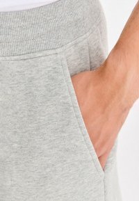 Ręka wsunięta do przedniej kieszeni jasnoszarych spodni dresowych z prążkowanym elastycznym pasem.