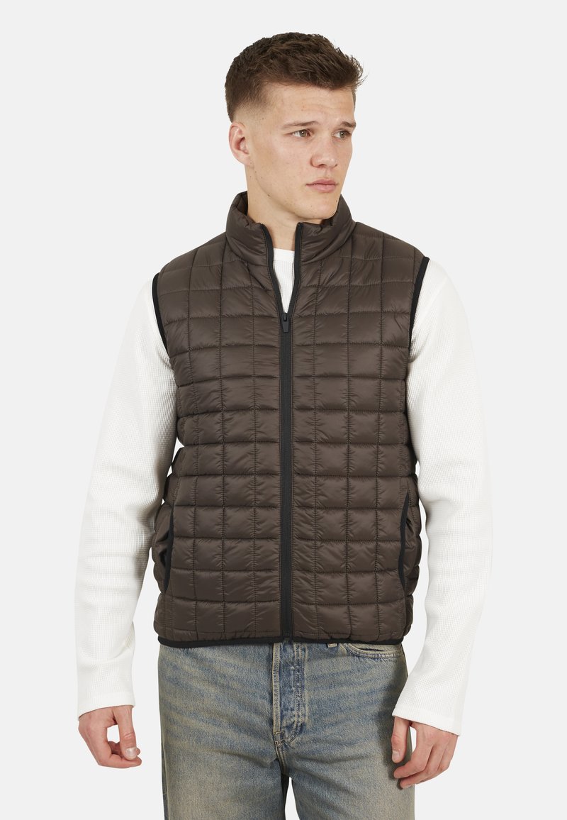 Gilet imbottito marrone scuro con colletto alto, zip frontale e tasche laterali, indossato sopra una camicia bianca a maniche lunghe.