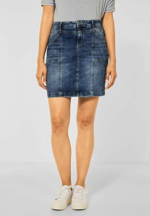 Femme portant une mini-jupe en jean délavé moyen avec des détails de couture à l'avant, associée à des baskets blanches à lacets et un haut gris clair rentré.