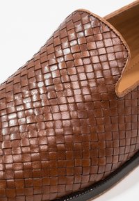 Chaussure slip-on en cuir marron avec texture tissée, finition brillante et forme arrondie. Comprend une semelle en caoutchouc noire contrastante.