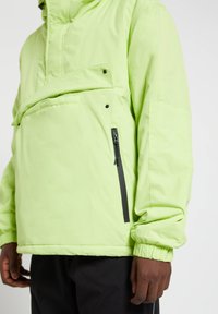 Hombre con abrigo anorak verde claro con bolsillo delantero de solapa y bolsillo negro con cremallera en el lateral, combinado con pantalones negros.