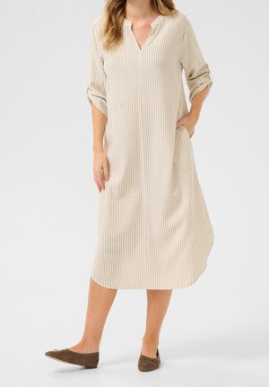 Frau trägt ein midi Kleid mit beigen und weißen vertikalen Streifen, dreiviertel hochgekrempelte Ärmel und braune flache Schuhe, steht mit einer Hand in der Tasche.