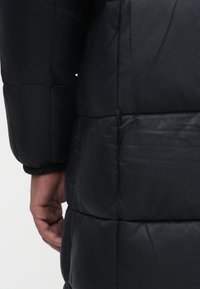 Giacca a vento nera con una superficie liscia e lucida. Il design include sezioni trapuntate orizzontali e polsini elasticizzati. Una mano è parzialmente visibile.