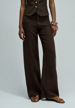 Persona que lleva pantalones anchos de color marrón oscuro, un chaleco a juego con botones dorados, sandalias y una pulsera gruesa en la muñeca derecha.